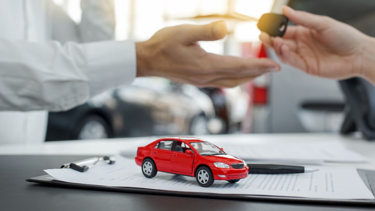 A man handing a car key to another man over a table with a red car toy on it