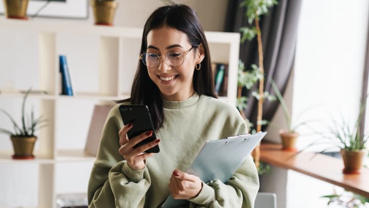 Dark haired woman holding a pile of documents whilst reading on her smartphone and smiling